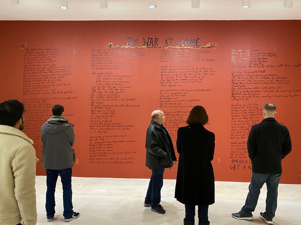 Five people stand in a gallery in front of a large red wall with black text  for The War At Home